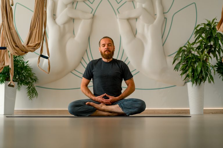A man sits cross-legged on a yoga mat, meditating in a studio adorned with large white hand sculptures and potted plants—an inspiring Sample Post capturing tranquility and mindfulness.
