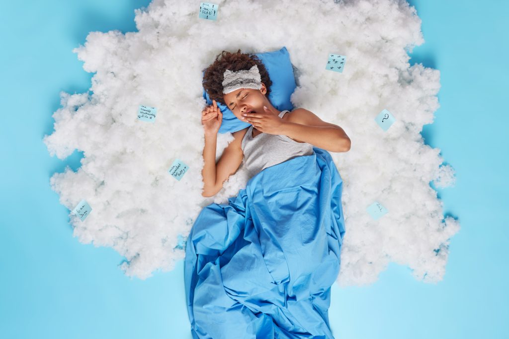 Person lying on blue bedding, surrounded by white clouds and small note cards, appearing to yawn or stretch with an eye mask on their forehead—your perfect Sample Post for relaxing vibes against a tranquil blue background.