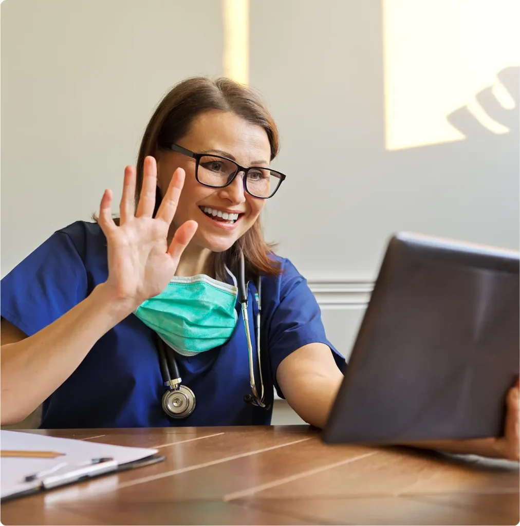 A healthcare professional in scrubs and a mask waves at a tablet screen while sitting at a desk.