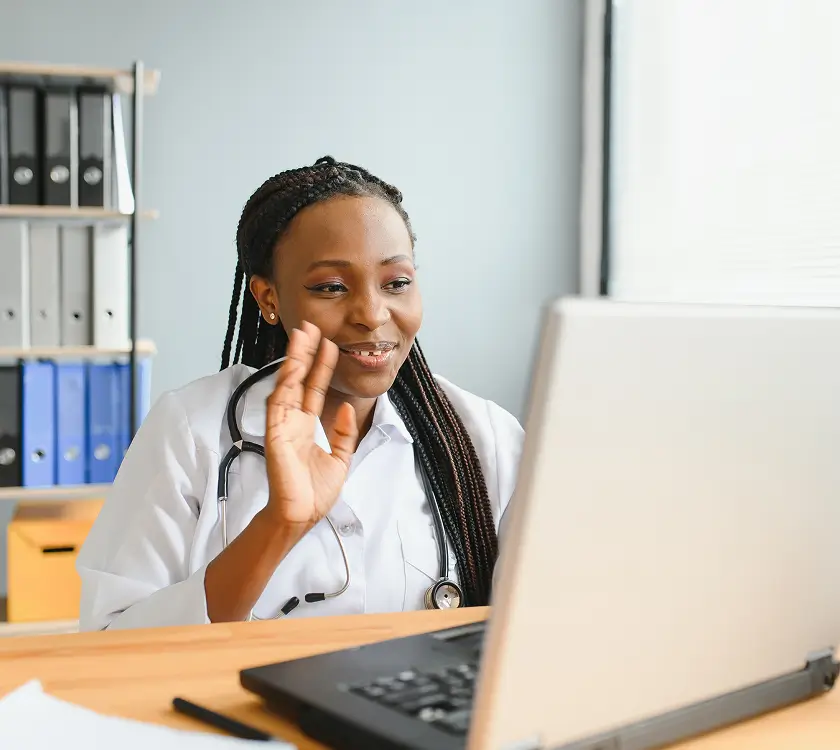 A person in a lab coat with a stethoscope sits at a desk, smiling and waving at a laptop. Shelves with binders are in the background.