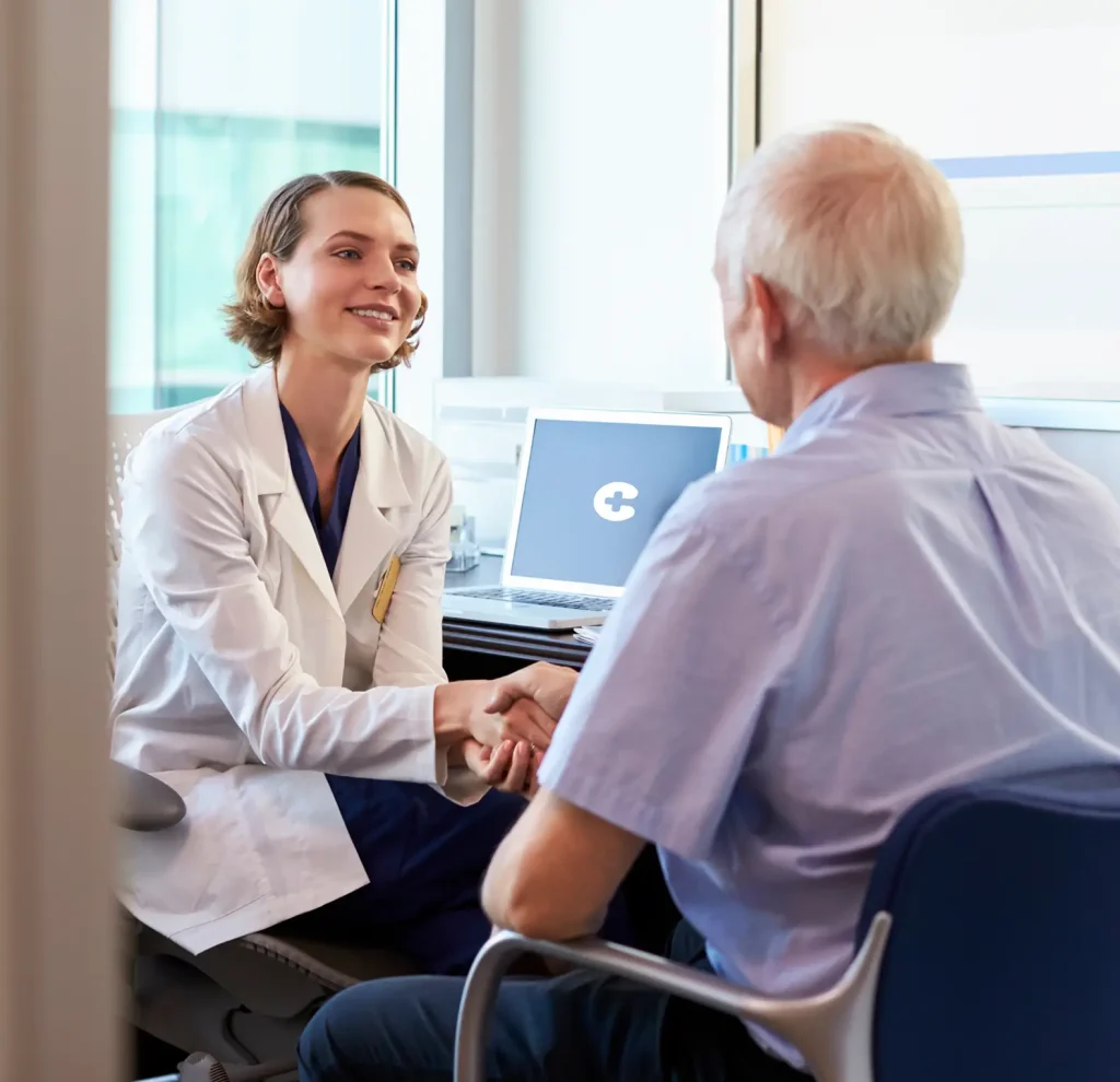 A doctor in a white coat shakes hands with an older man in an office setting, with a laptop on the desk nearby.